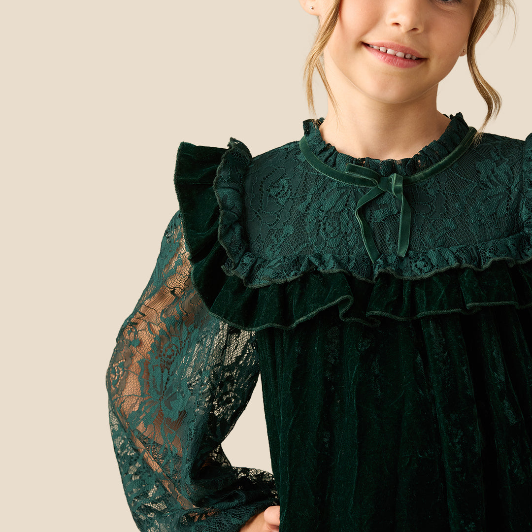 A young girl wearing a dark forest green velvet dress with lace sleeves and a ruffled high neckline, photographed against a solid beige background.
