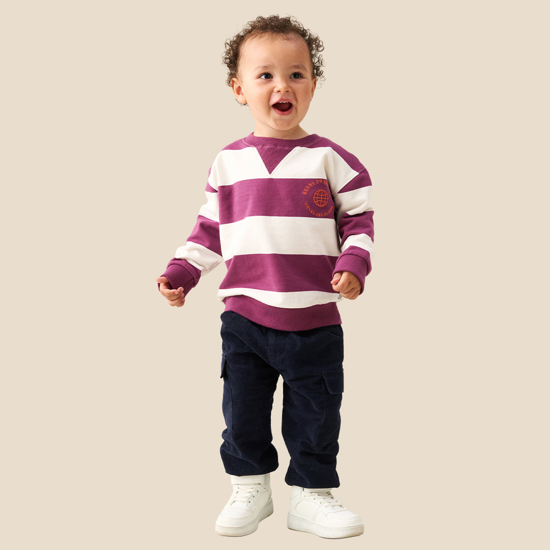 A toddler standing against a solid beige background, wearing a purple and white horizontally striped long-sleeve sweatshirt, dark navy pants, and white sneakers.