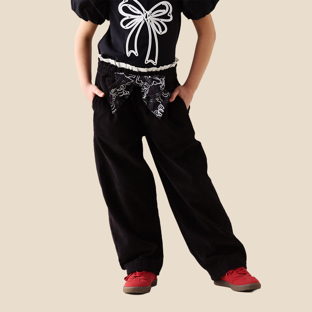 A child wearing black wide-leg pants with a white bow and floral pattern at the waistband, paired with a black top and red sneakers, standing against a solid beige background.