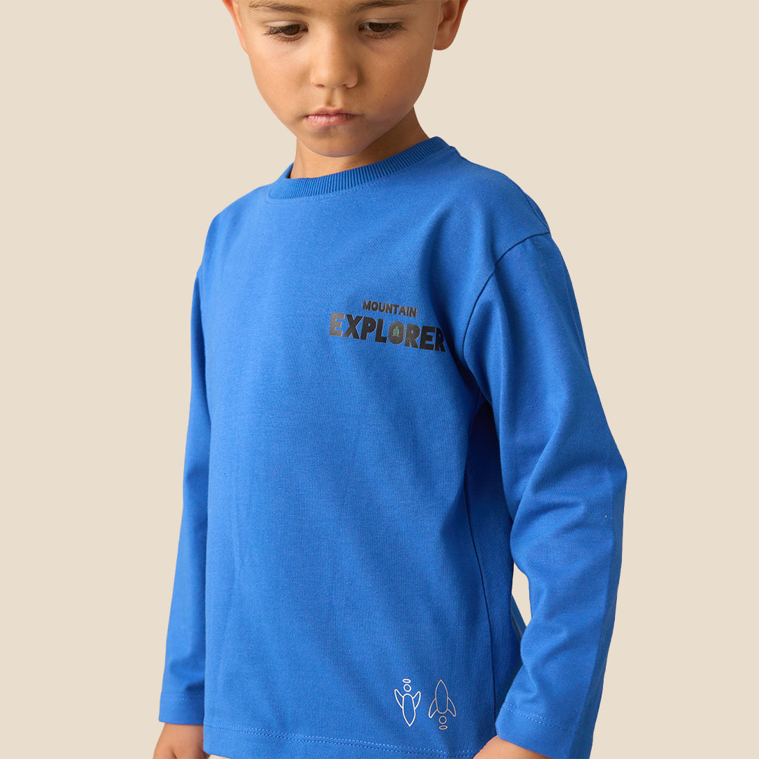 A young boy modeling a two-piece pajama set consisting of a bright blue long-sleeve shirt and navy blue wide-leg pants with pink text on the leg, set against a solid beige background.