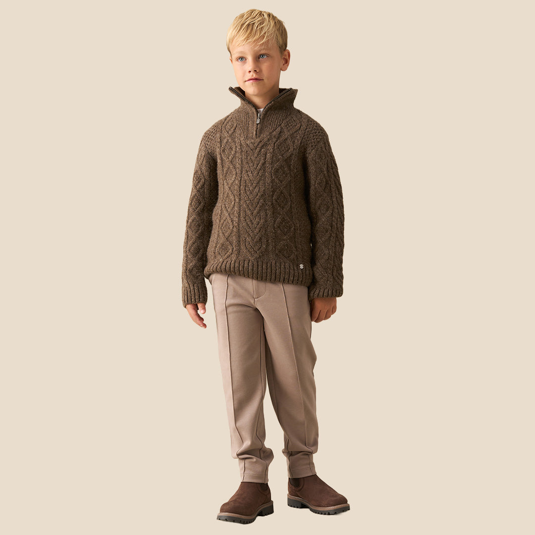 A child wearing a brown cable-knit mock-neck sweater and light pink trousers, standing indoors near a white wall and window. Another child in a brown-and-white plaid shirt is partially visible in the foreground.
