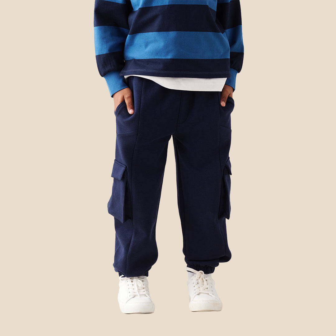 A child wearing navy blue cargo pants with side flap pockets, a blue and navy striped long-sleeve shirt, and white sneakers, standing against a solid beige background.