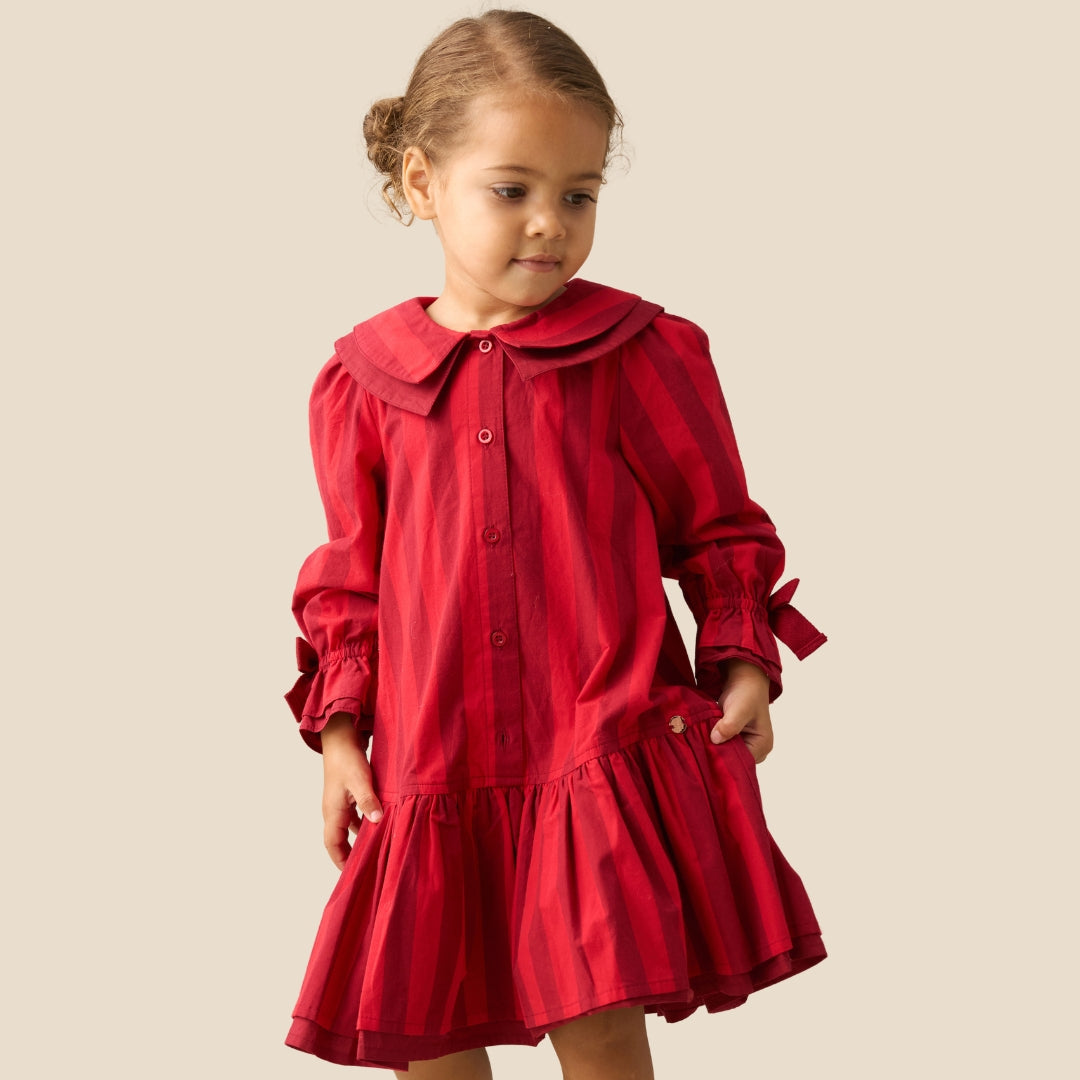 A young girl with her hair in a bun wearing a red long-sleeve collared dress with a button-up front and ruffled skirt, standing against a solid beige background.