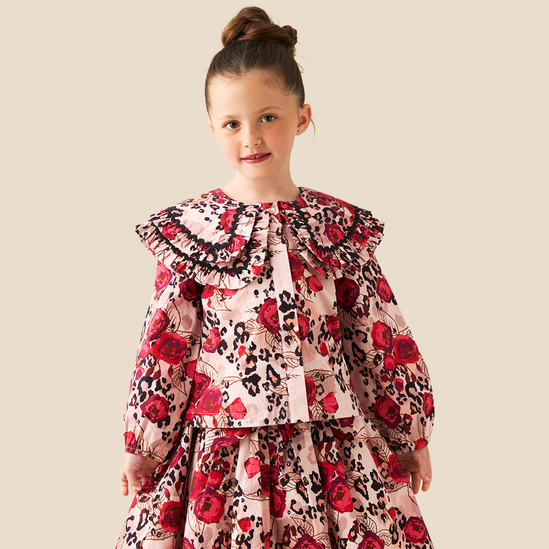 A young girl with her hair in a bun stands wearing a long-sleeve dress with a red rose and black leopard print pattern on a light pink background, set against a solid beige studio backdrop.