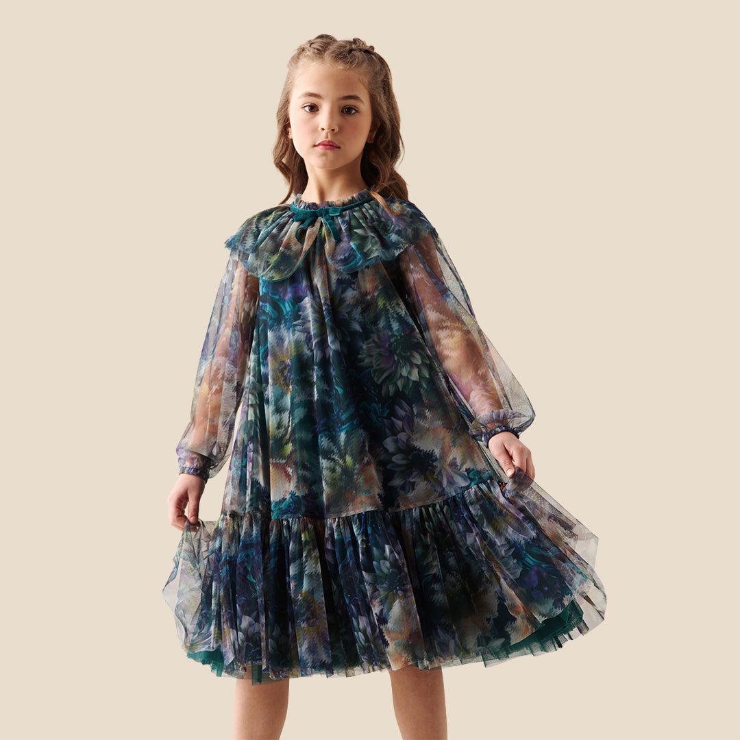 A young girl modeling a sheer, tiered tulle dress with a large ruffled collar and a blue, green, and purple floral watercolor pattern, set against a solid beige background.
