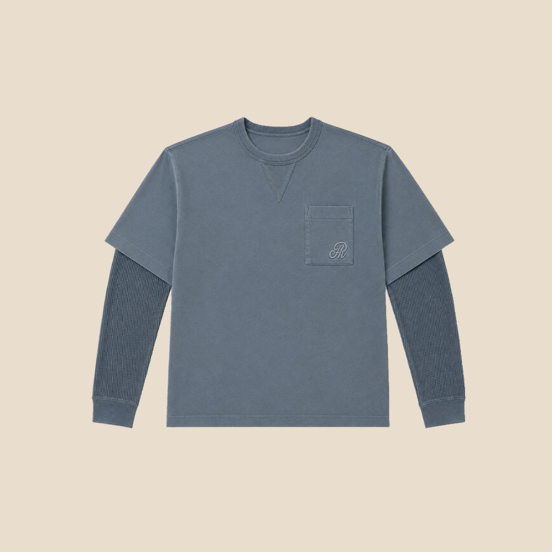 A young boy modeling a washed slate blue long-sleeve crewneck shirt with a chest pocket, paired with beige cargo pants, against a solid beige background.