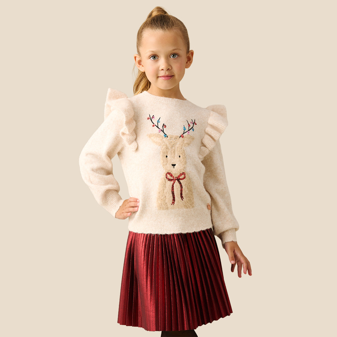 A young girl with blonde hair in a ponytail models a cream-colored knit sweater featuring a large embroidered reindeer on the front with colorful antlers and a red bow. She is wearing a shiny red skirt against a solid light beige background.
