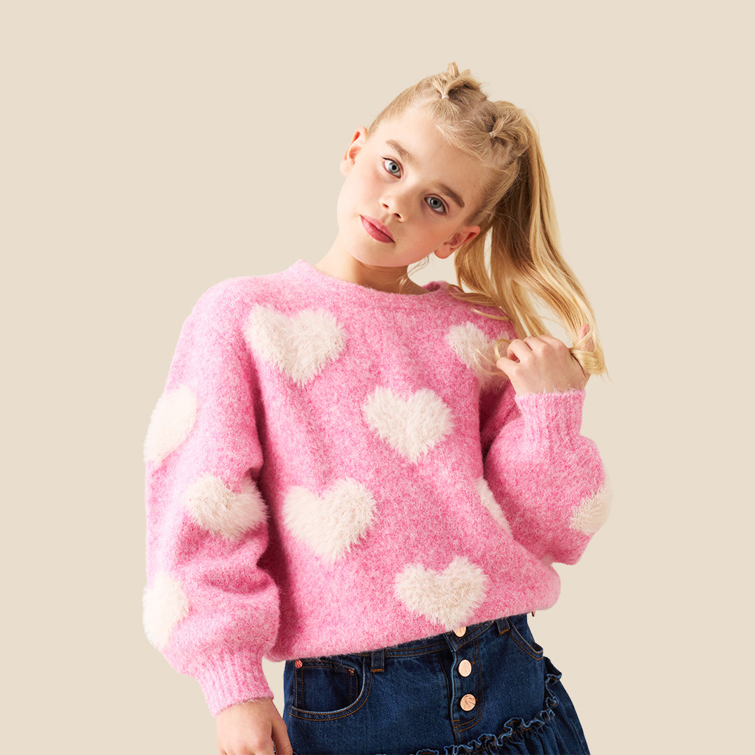 A young girl with blonde hair styled in buns wears a pink fuzzy knit sweater with fluffy white heart details, posing with her hands forming a heart shape in front of her chest. She is wearing a dark blue denim skirt.