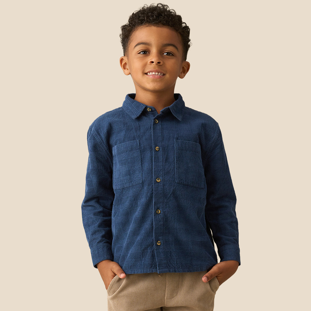 A young boy with curly dark hair wearing a dark blue long-sleeve button-up shirt and khaki pants, standing against a solid beige background.