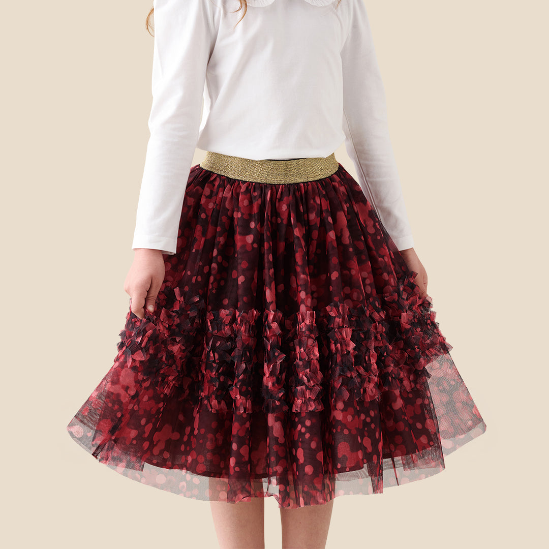 A child wearing a white long-sleeve top and a tiered tulle skirt in dark red and black floral print with a gold waistband, shown against a plain beige background.
