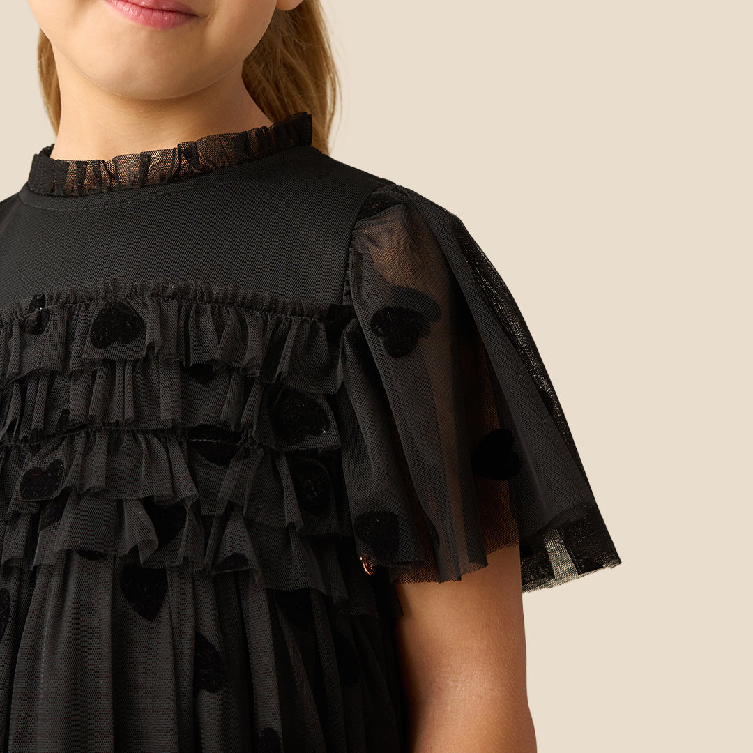 A young girl with her hair in a ponytail models a black tulle dress with heart patterns and sheer flutter sleeves, set against a solid beige background.
