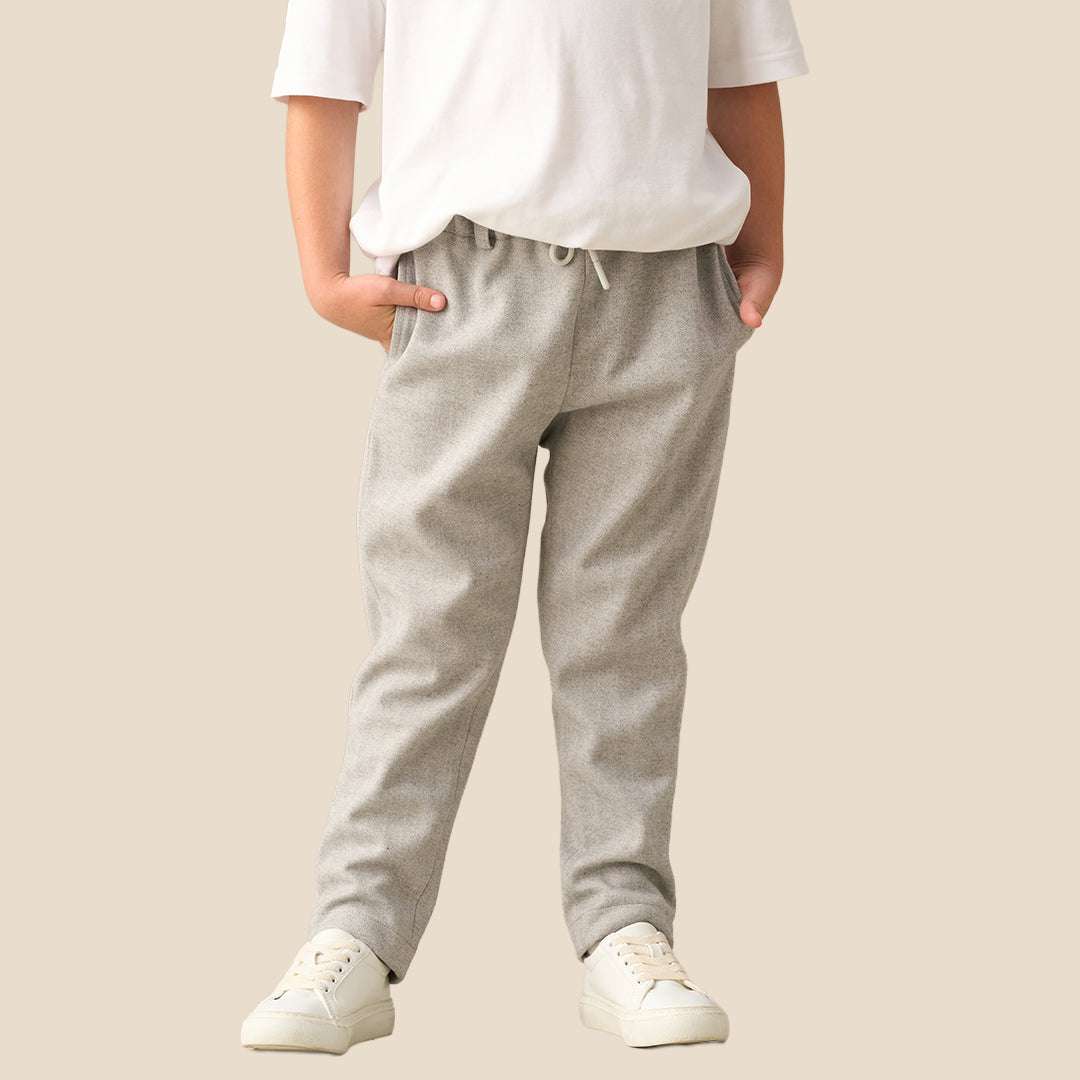 A child wearing light gray heathered sweatpants with a white t-shirt and white sneakers, standing against a solid beige background.