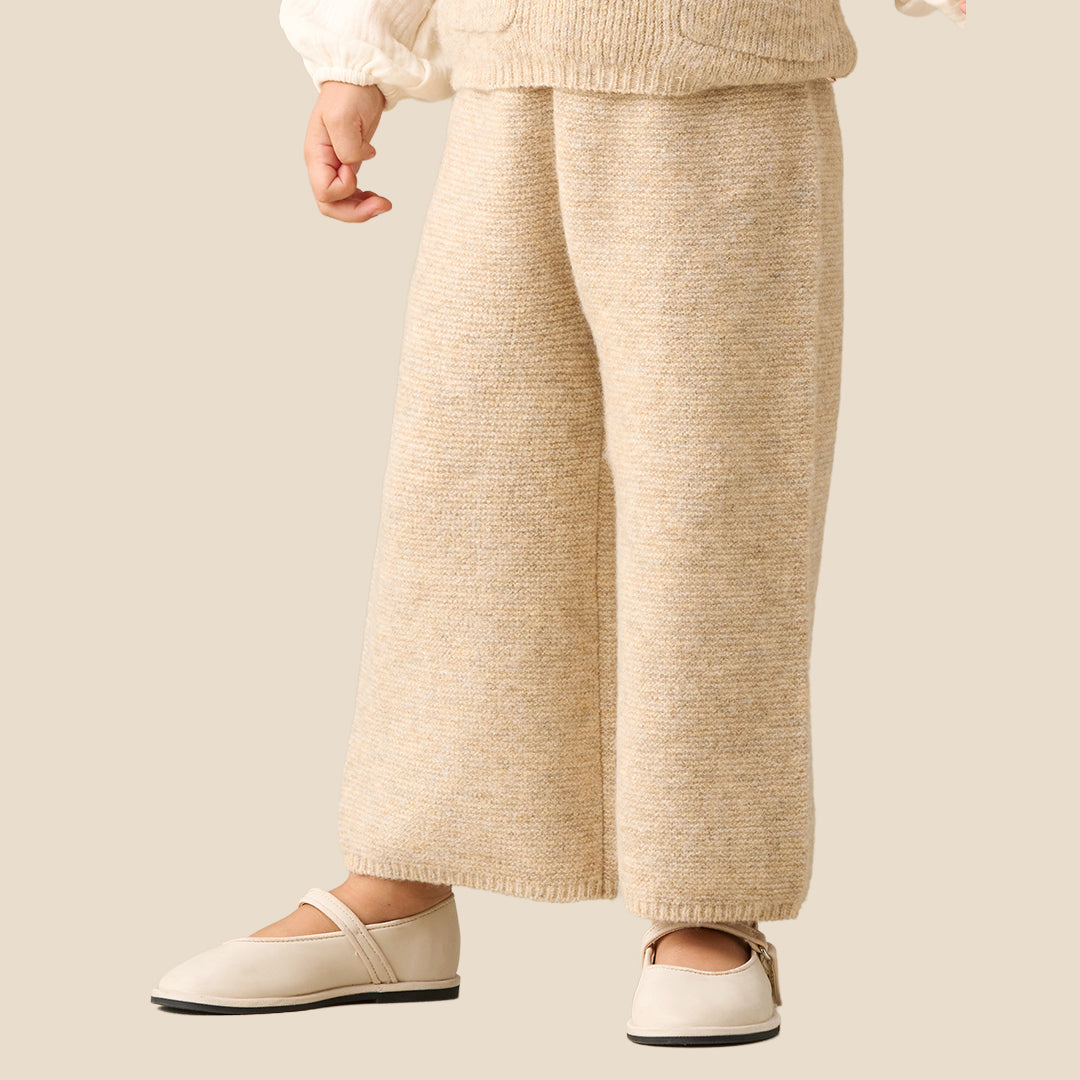 A young child modeling a cream and beige knit outfit, consisting of a long-sleeve top with a ruffled collar and matching knit vest and pants, set against a solid light beige background.