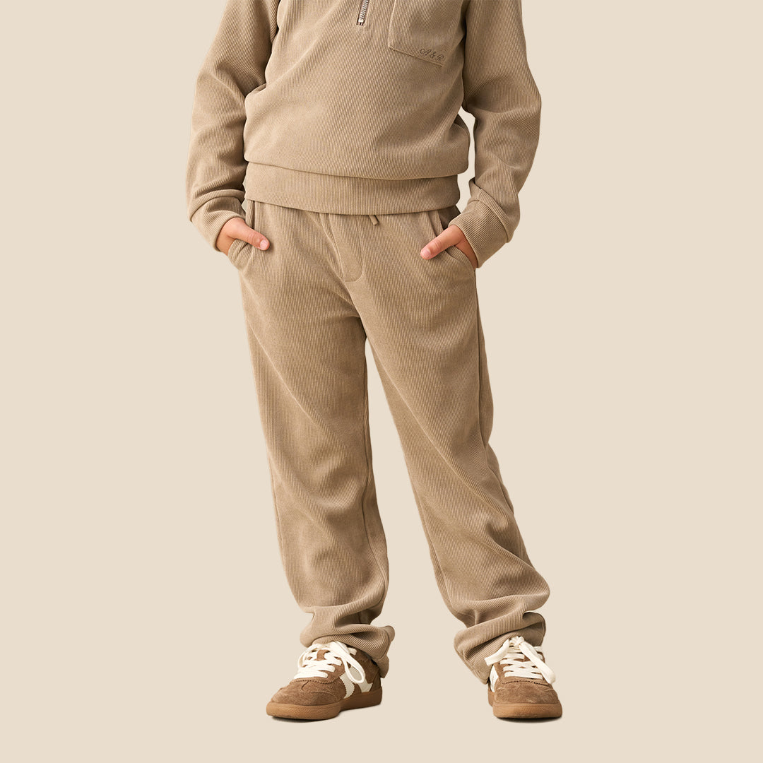 A child wearing a matching beige fleece tracksuit with a half-zip sweatshirt and relaxed-fit pants, paired with brown sneakers, against a solid light beige background.