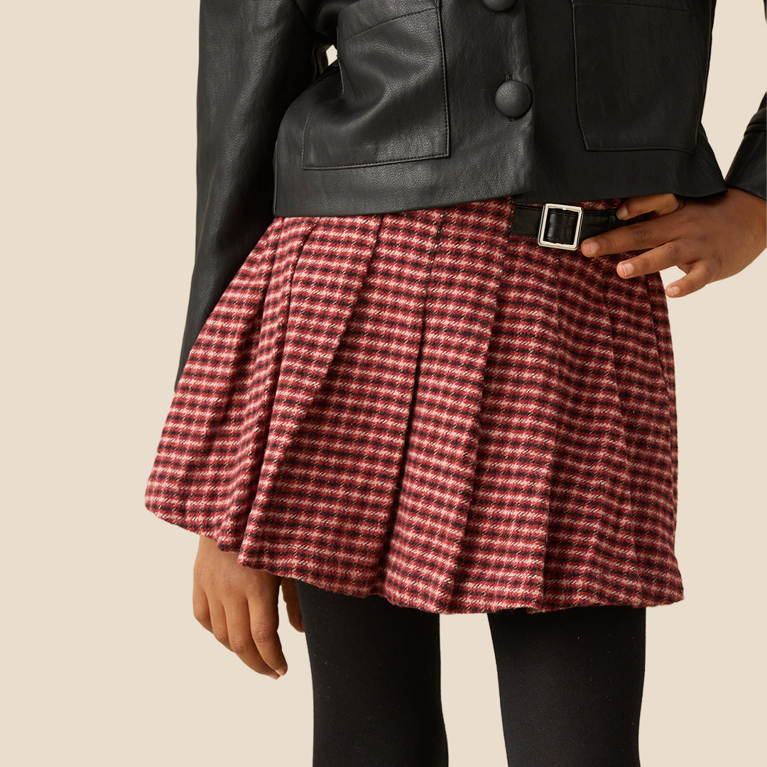 A close-up of a pleated skirt in a red and black gingham pattern, styled with a black leather jacket and black tights against a solid beige background.