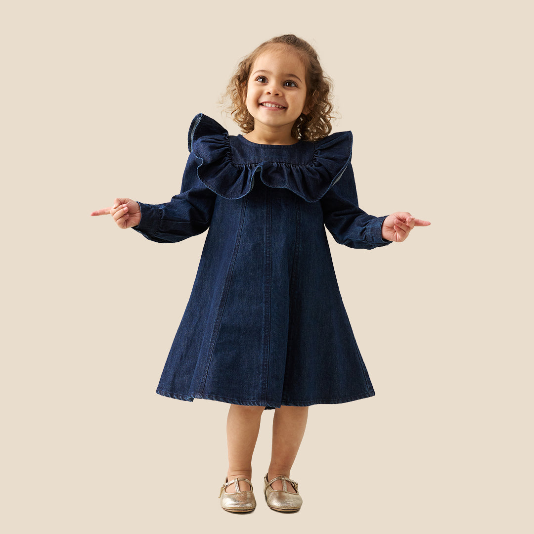 A young child wearing a dark blue denim dress with dramatic ruffled shoulders, standing against a solid beige background.