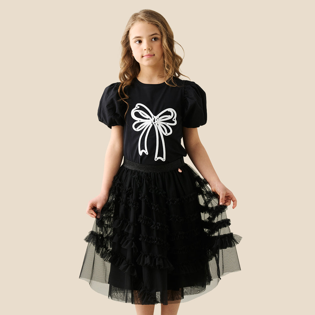 A child wearing a black puff-sleeve top with a large white bow graphic and a layered black ruffled tulle skirt, shown against a plain beige studio background.