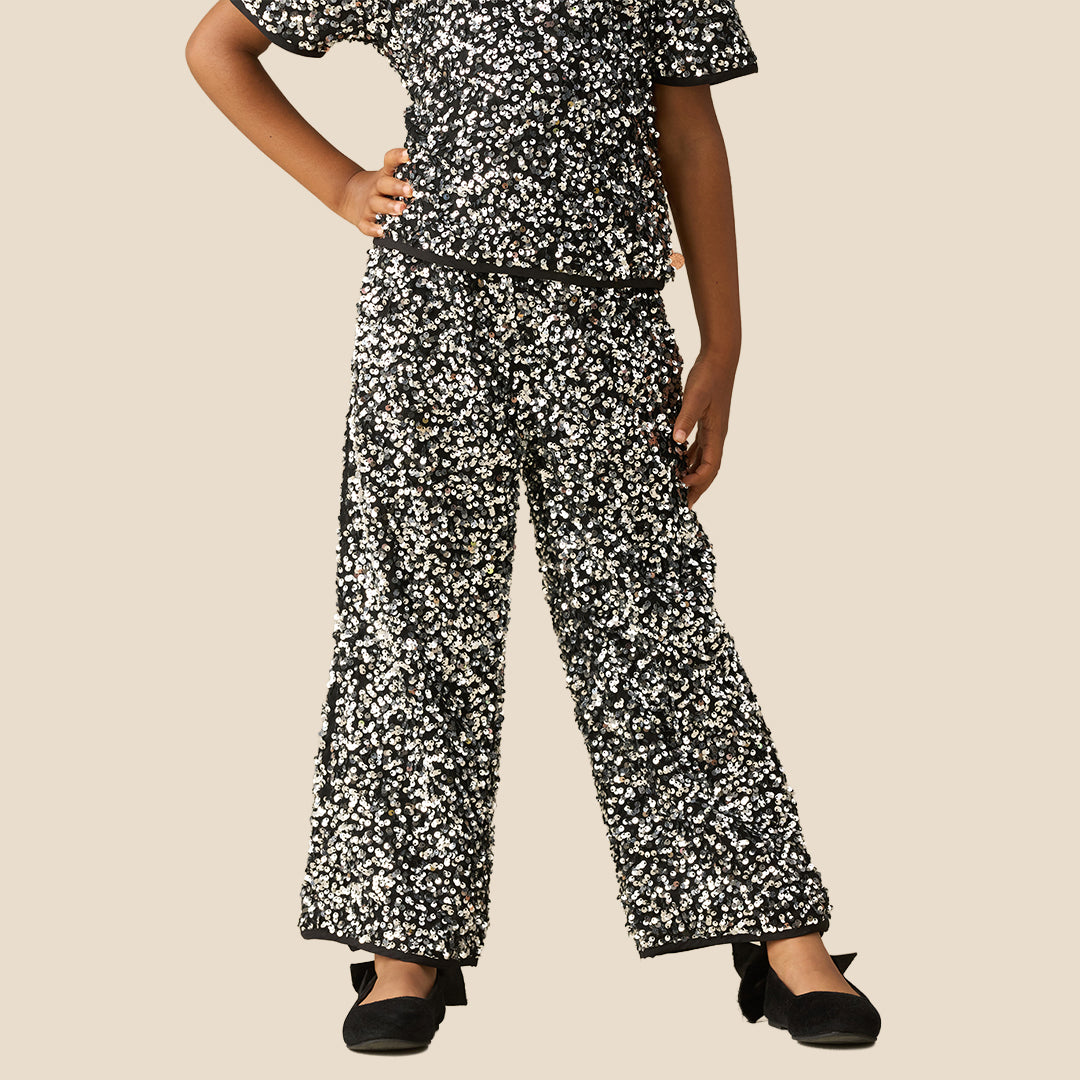 A child modeling a matching two-piece outfit with a black and white speckled pattern, featuring a short-sleeve top and wide-leg pants, styled with black flat shoes against a solid beige background.