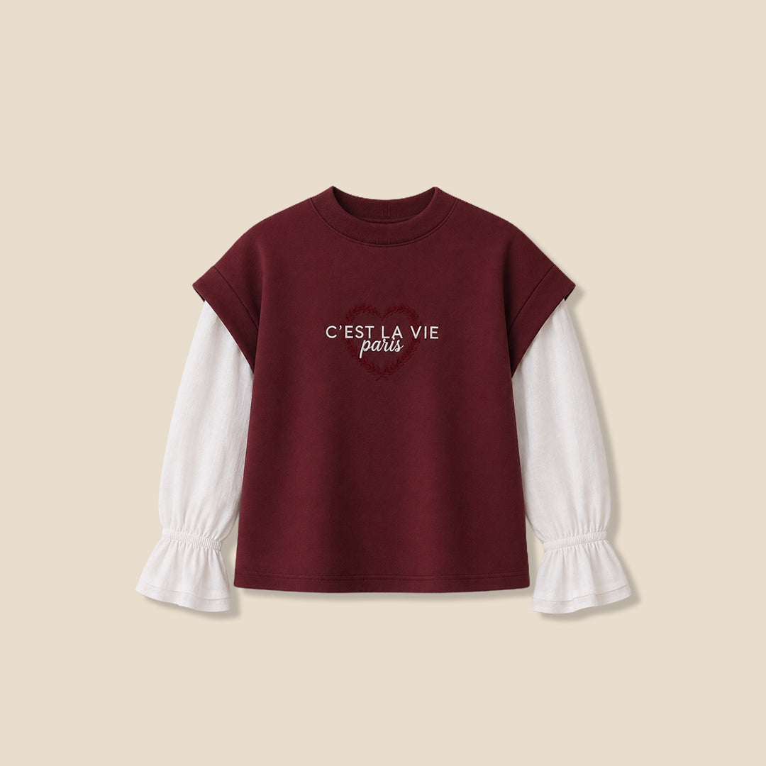 A young girl with blonde pigtails wearing a maroon cropped t-shirt with white sleeves and 'C'EST LA VIE paris' printed in white, layered over a white long-sleeve blouse with ruffled cuffs, and blue jeans, against a solid beige background.