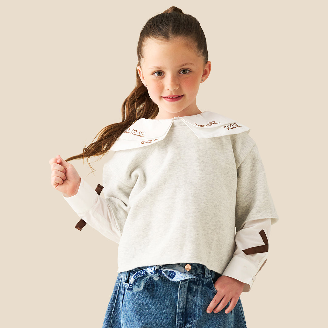A young girl with her hair in a ponytail models a light gray long-sleeve top featuring a large white collar with brown embroidered details and matching brown accents on the sleeves, paired with blue jeans against a solid beige background.