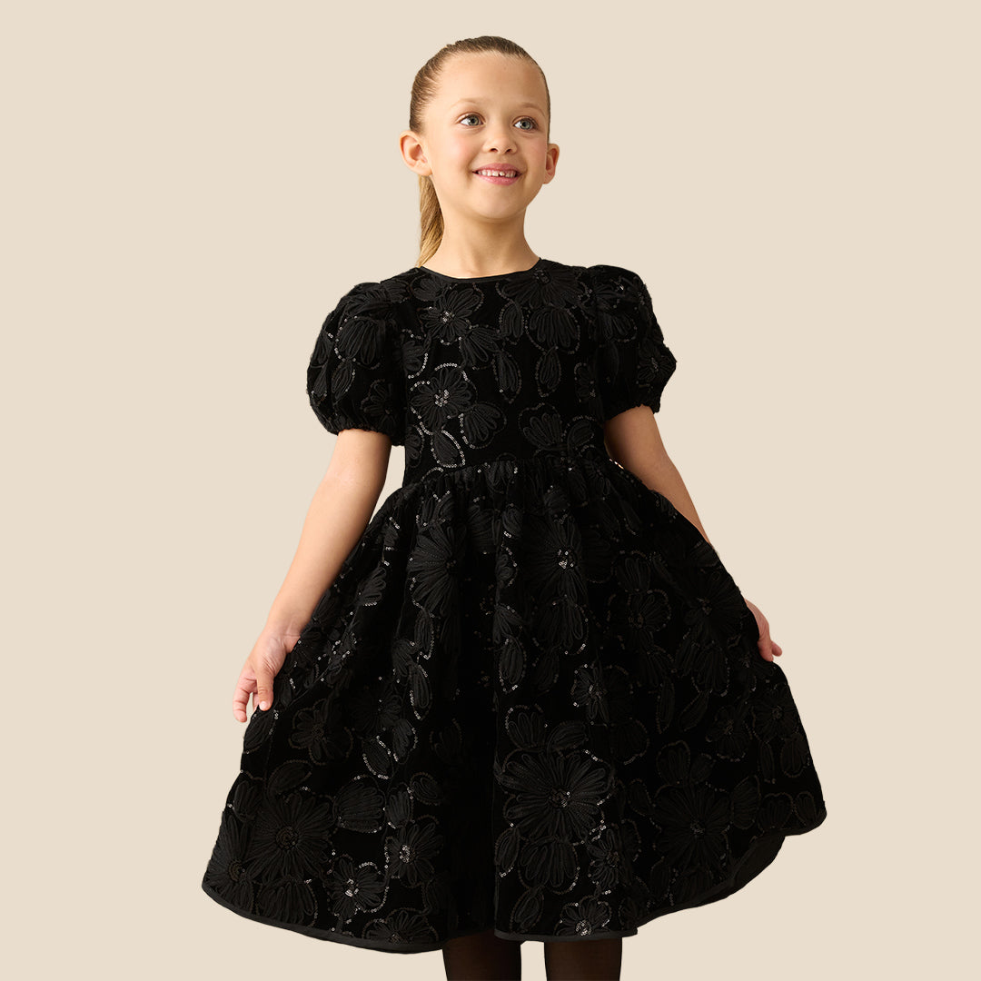 A young girl with her hair in a ponytail models a black short-sleeve dress with textured floral applique, standing against a solid beige background.