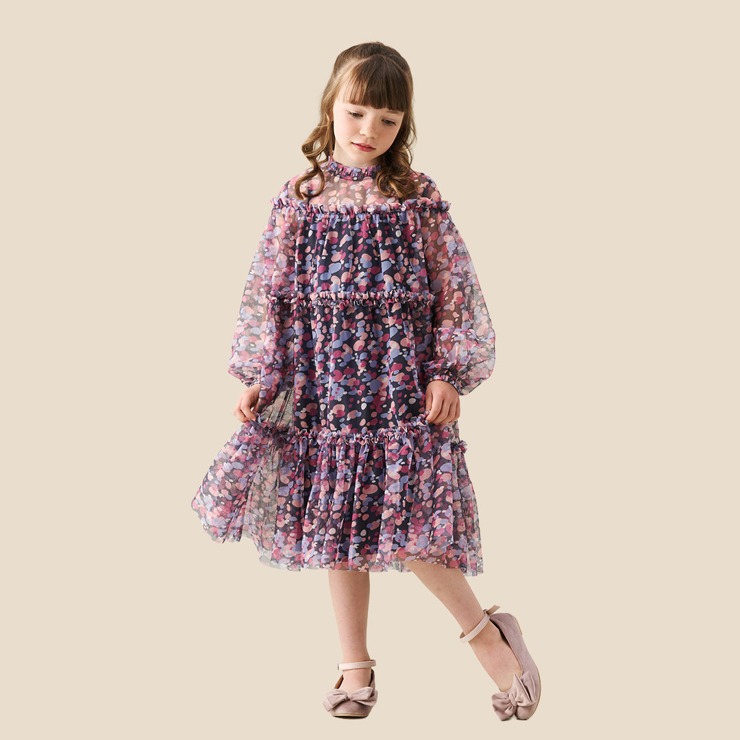 A young girl with curly hair stands on a checkered porch wearing a long-sleeved tiered dress with a pink, purple, and blue floral pattern. She is posing behind a black wrought-iron fence.