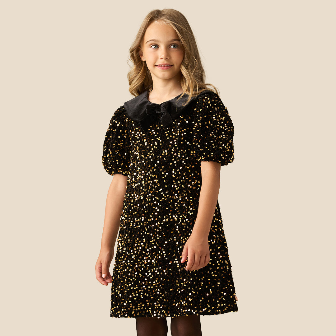 A young girl with blonde hair wearing a black short-sleeve dress with gold sequins and a black ruffled Peter Pan collar, standing against a solid beige background.