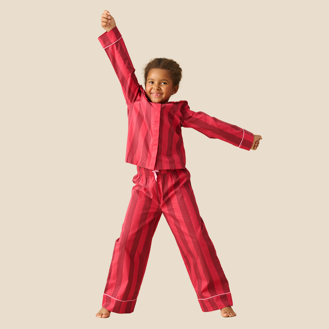 A young girl modeling a two-piece pajama set with vertical pink and red stripes, featuring a ruffled collar and white piping details, set against a solid beige background.