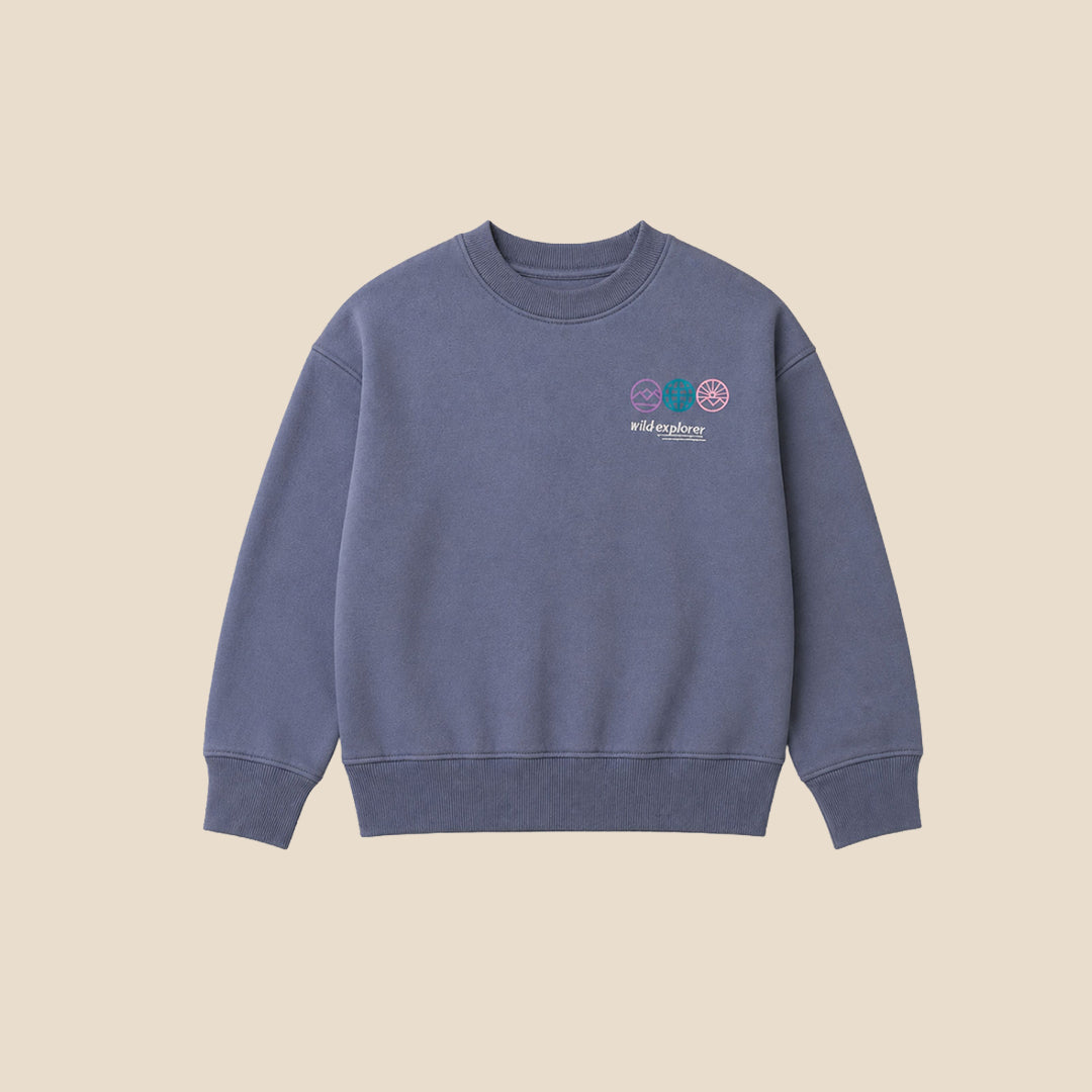 A young boy wearing a slate blue crewneck sweatshirt with a small colorful graphic on the left chest, paired with gray jeans, standing against a solid beige background.