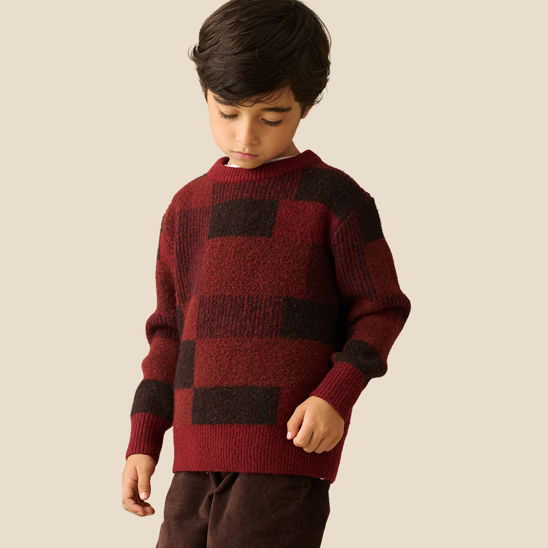 A young boy wearing a textured knit sweater with a dark red and black checkered pattern, paired with dark brown pants, set against a solid light beige background.