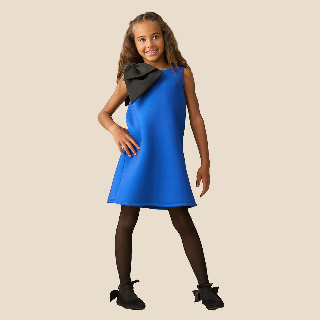 A young girl with long brown hair wearing a bright blue sleeveless dress with a large black bow on the shoulder, standing against a solid beige background.