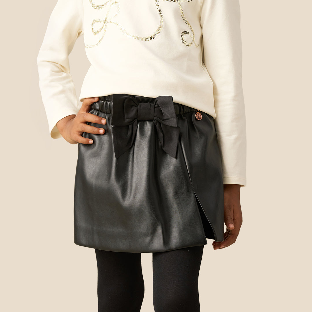 A child wearing a black faux-leather wrap mini skirt with a large black fabric bow at the waist, styled with a cream long-sleeve top and black tights against a solid beige background.