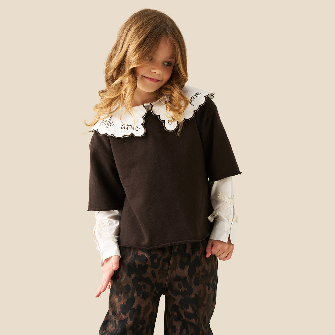 A young girl with wavy light brown hair is wearing a dark brown short-sleeve top with a large white ruffled collar featuring black cursive embroidery, layered over a white long-sleeve shirt, paired with brown and black leopard-print pants, against a solid beige background.
