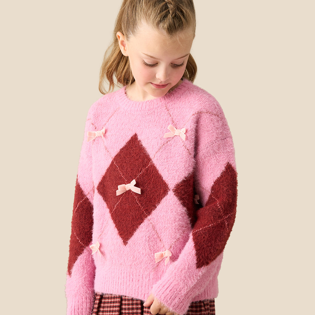 A young girl modeling a pink and dark red argyle sweater with small bow details, paired with a red and black gingham pleated skirt, against a solid beige background.