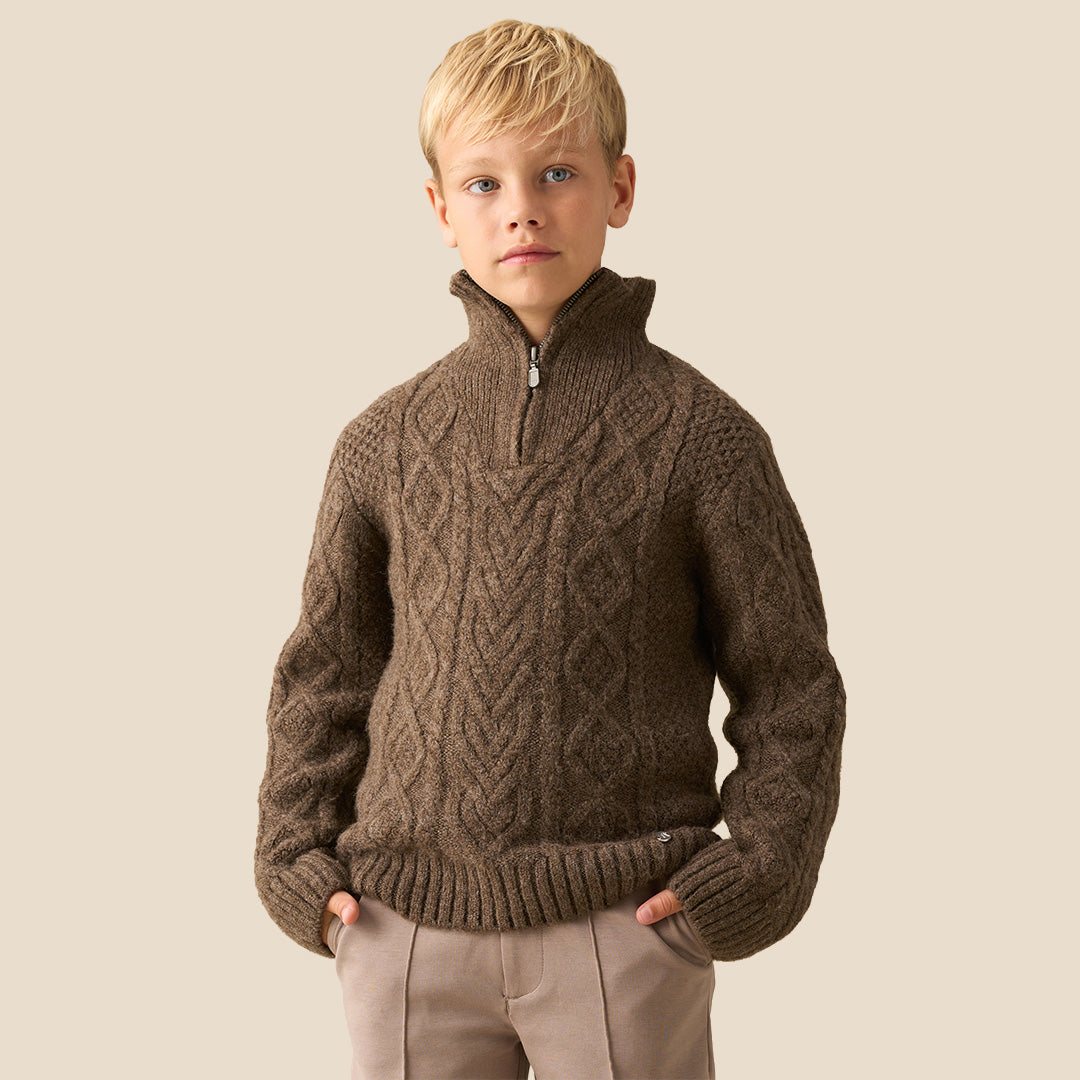 A child wearing a brown cable-knit mock-neck sweater and light pink trousers, standing indoors near a white wall and window. Another child in a brown-and-white plaid shirt is partially visible in the foreground.
