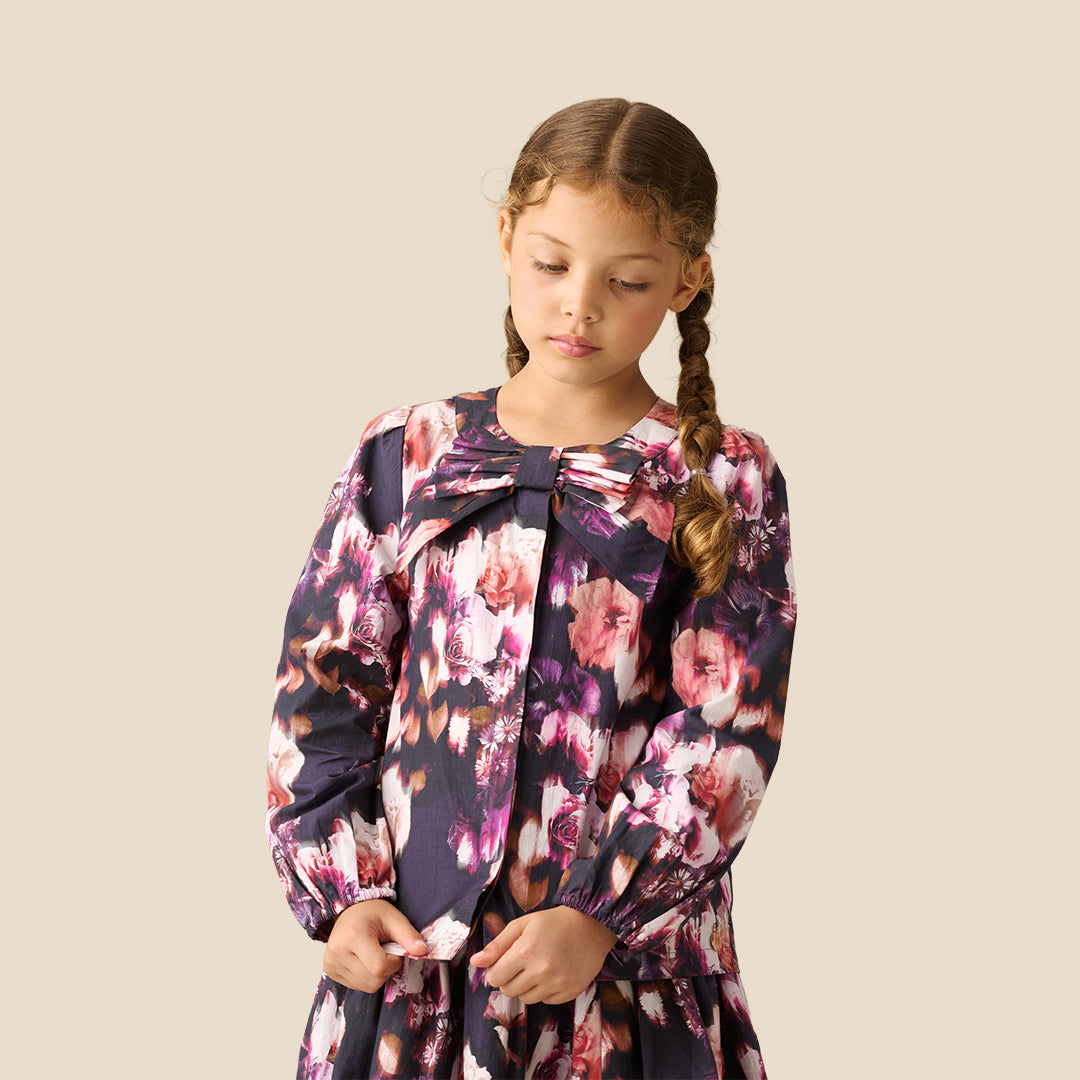 A young girl with braided hair wearing a long-sleeve floral blouse with a large bow at the collar, photographed against a plain light beige studio background.