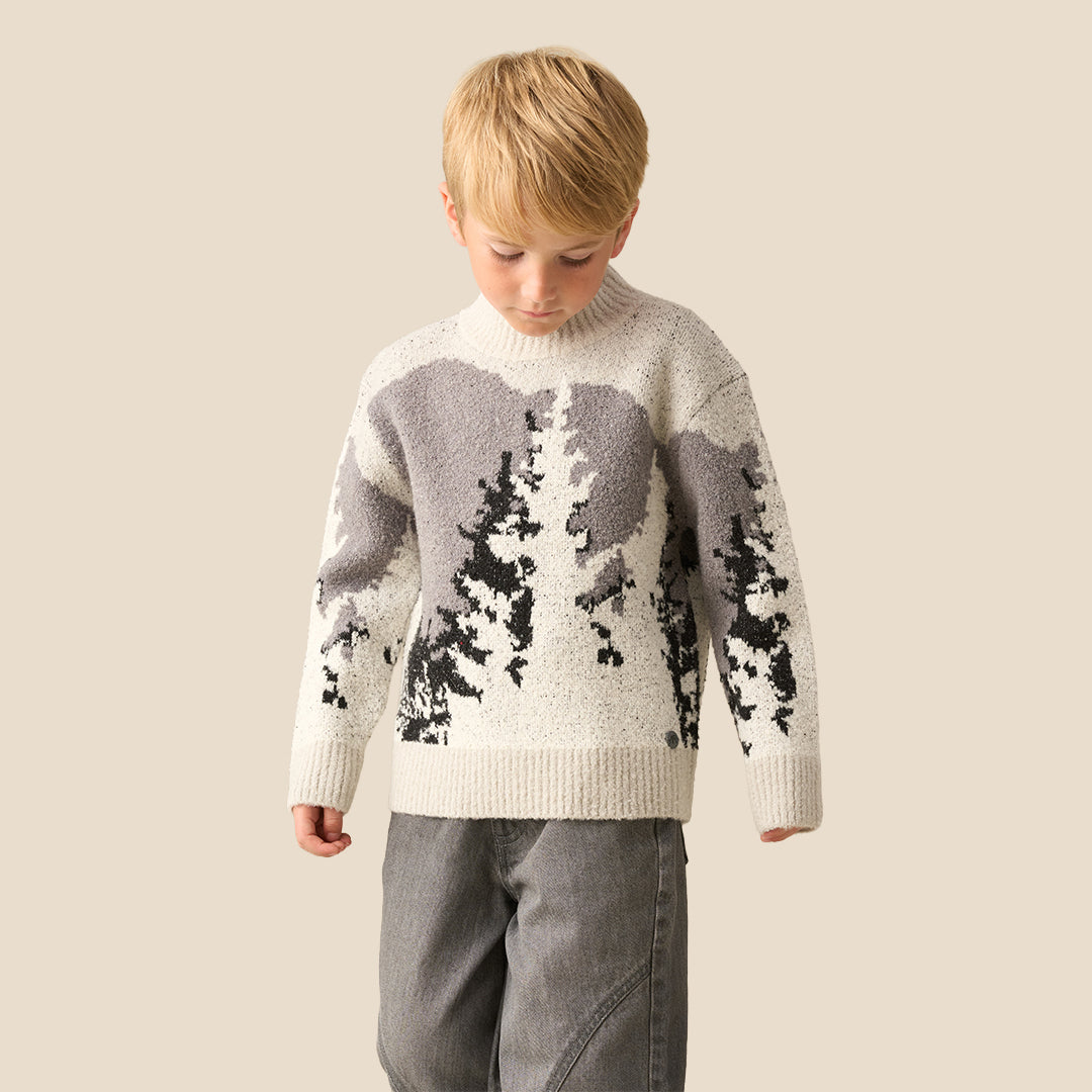 A young boy with blonde hair wearing a knit mock-neck sweater with a gray and black tree pattern, standing against a solid beige background.