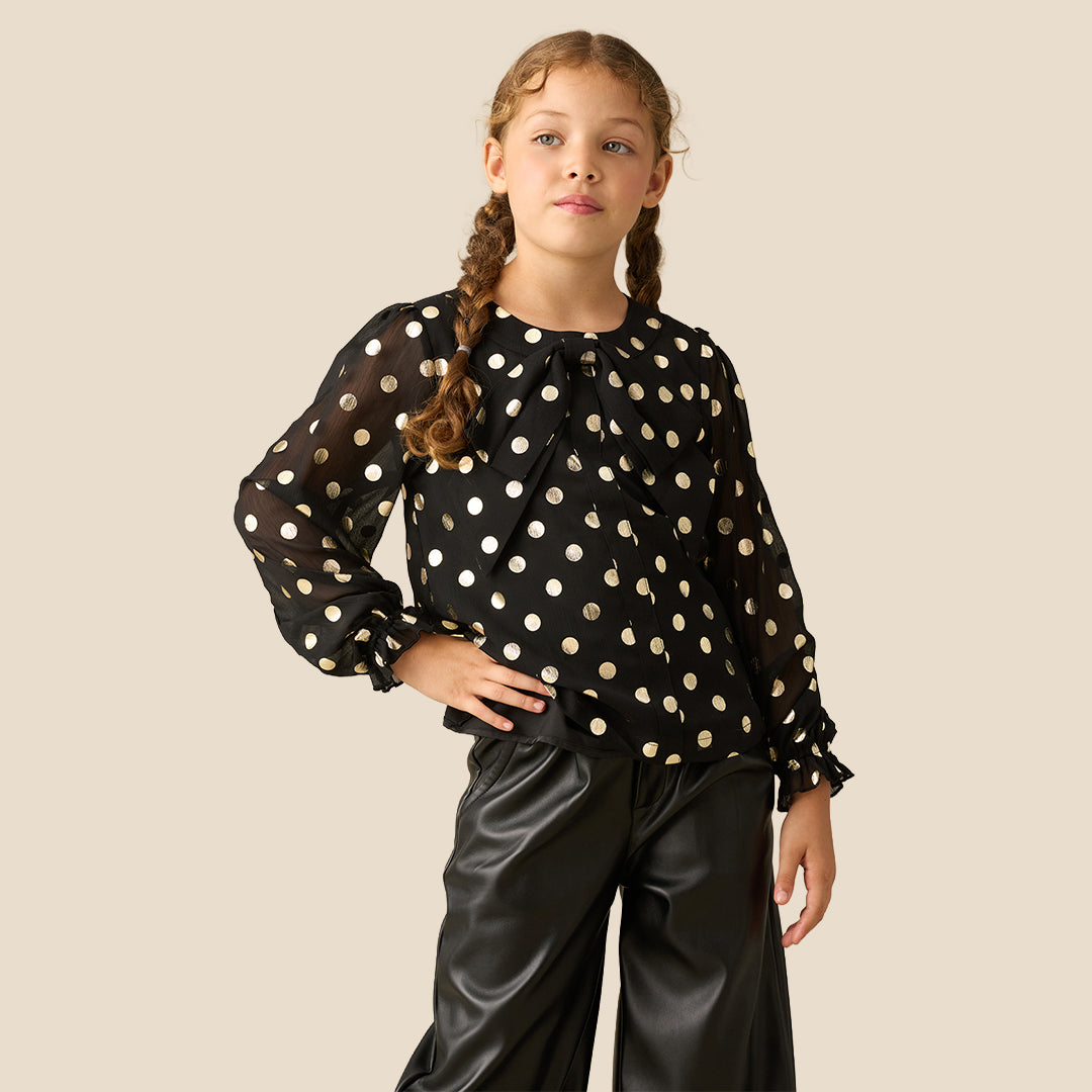A young girl with braided hair wearing a black long-sleeve blouse with gold polka dots and black faux-leather pants, standing against a solid beige background.