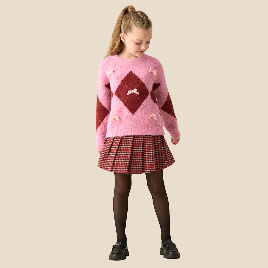 A young girl modeling a pink and dark red argyle sweater with small bow details, paired with a red and black gingham pleated skirt, against a solid beige background.