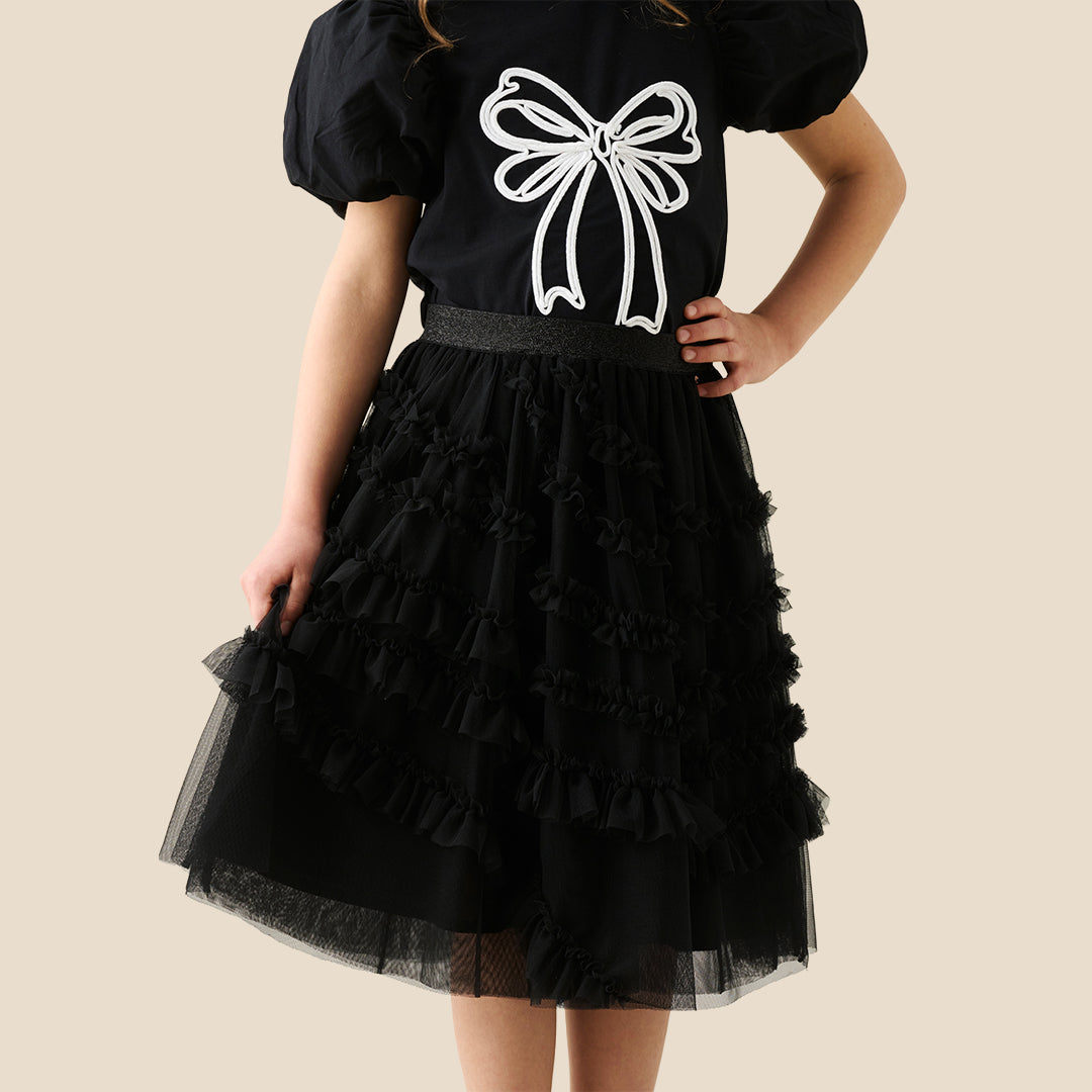 A child wearing a black puff-sleeve top with a large white bow graphic and a layered black ruffled tulle skirt, shown against a plain beige studio background.