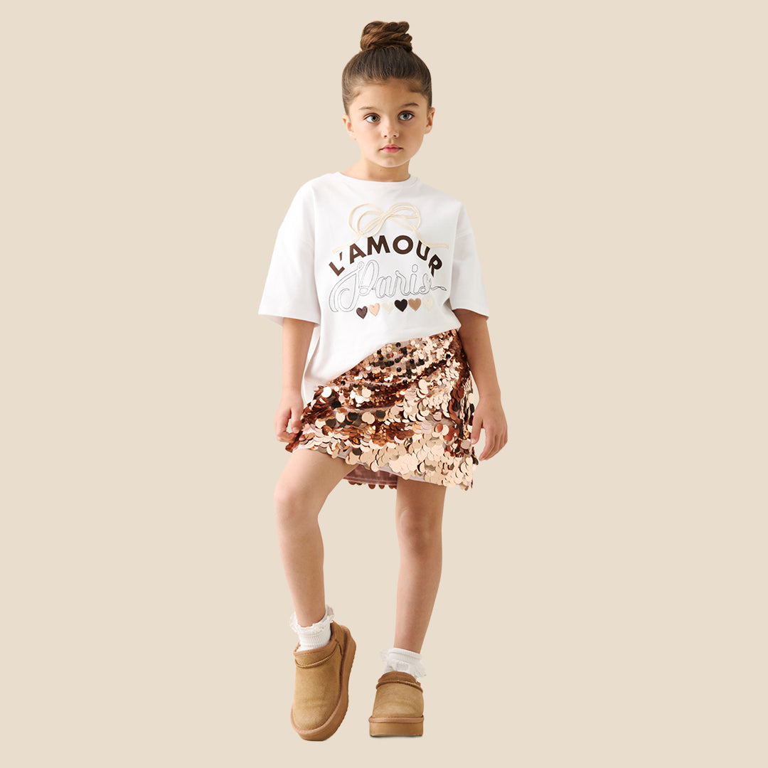 A child wearing a white graphic T-shirt and a short skirt covered in large, reversible sequins that shift between rose gold and bronze, photographed against a plain beige studio background.