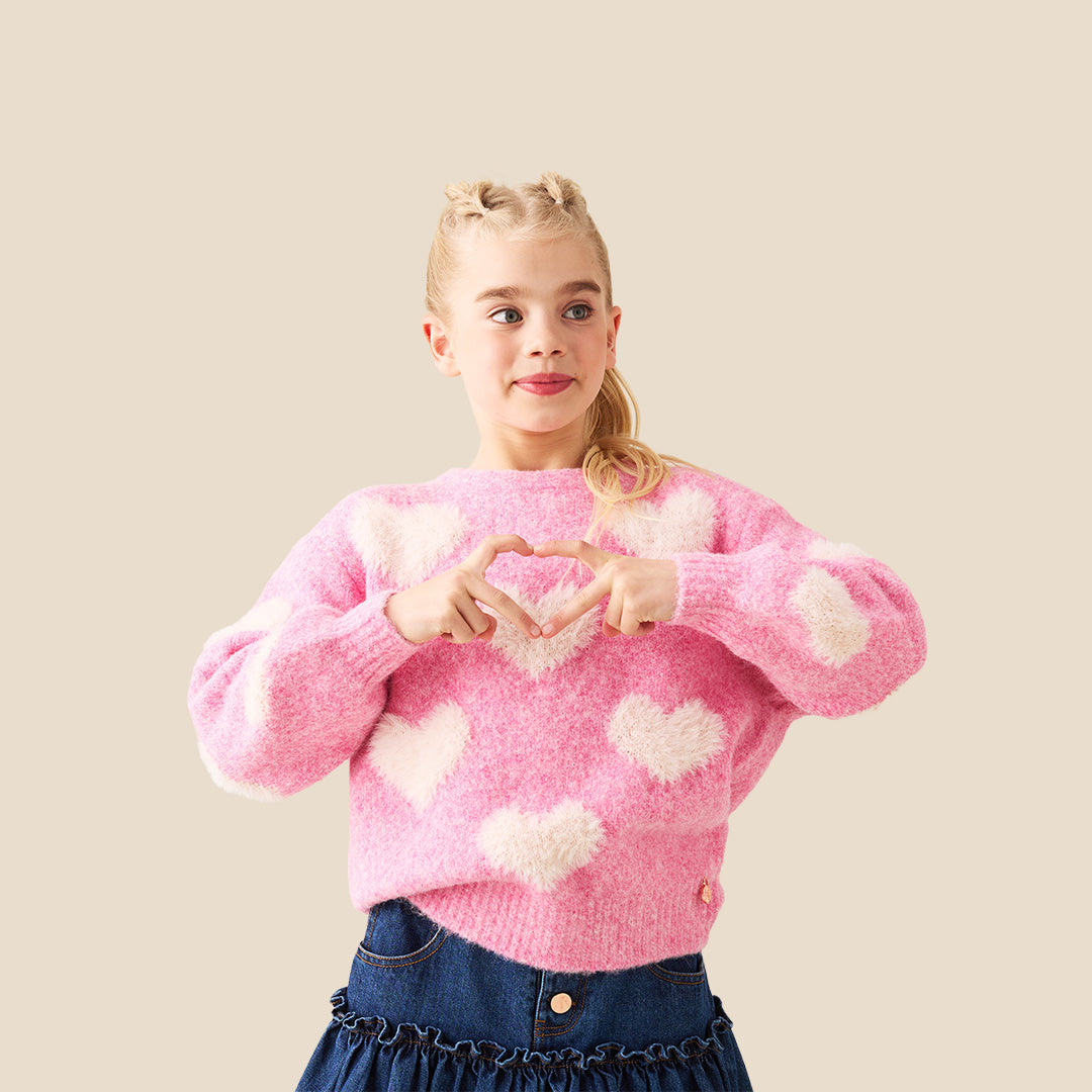 A young girl with blonde hair styled in buns wears a pink fuzzy knit sweater with fluffy white heart details, posing with her hands forming a heart shape in front of her chest. She is wearing a dark blue denim skirt.