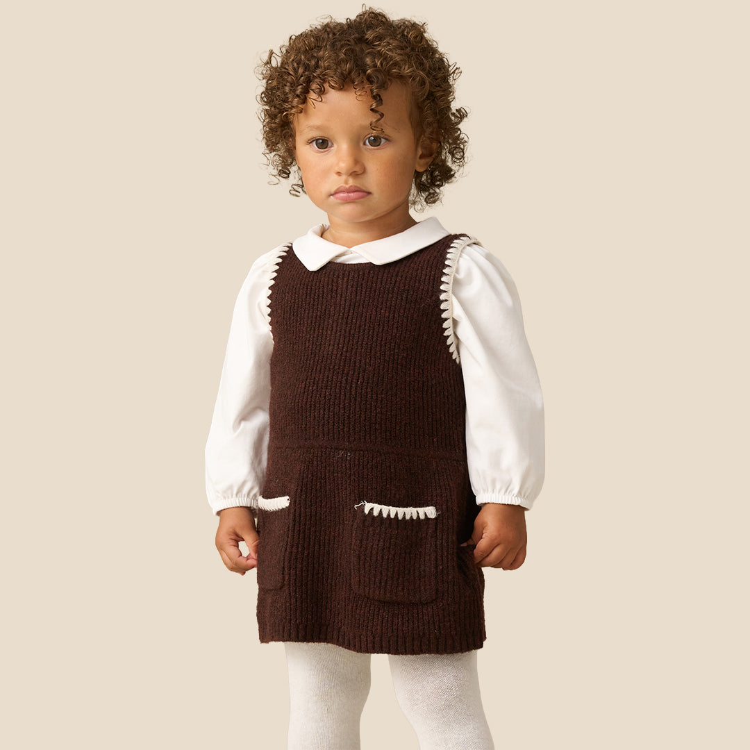 A toddler with curly brown hair wearing a dark brown corduroy pinafore dress over a white long-sleeve collared shirt and white tights, standing against a solid light beige background.