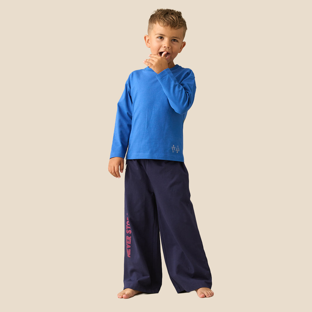 A young boy modeling a two-piece pajama set consisting of a bright blue long-sleeve shirt and navy blue wide-leg pants with pink text on the leg, set against a solid beige background.