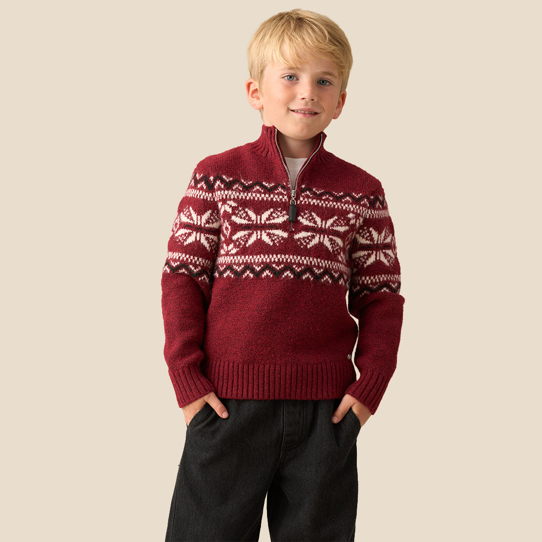 A young boy smiling and wearing a burgundy knit quarter-zip sweater with a white and dark red Fair Isle snowflake pattern across the chest and shoulders, set against a solid beige background.