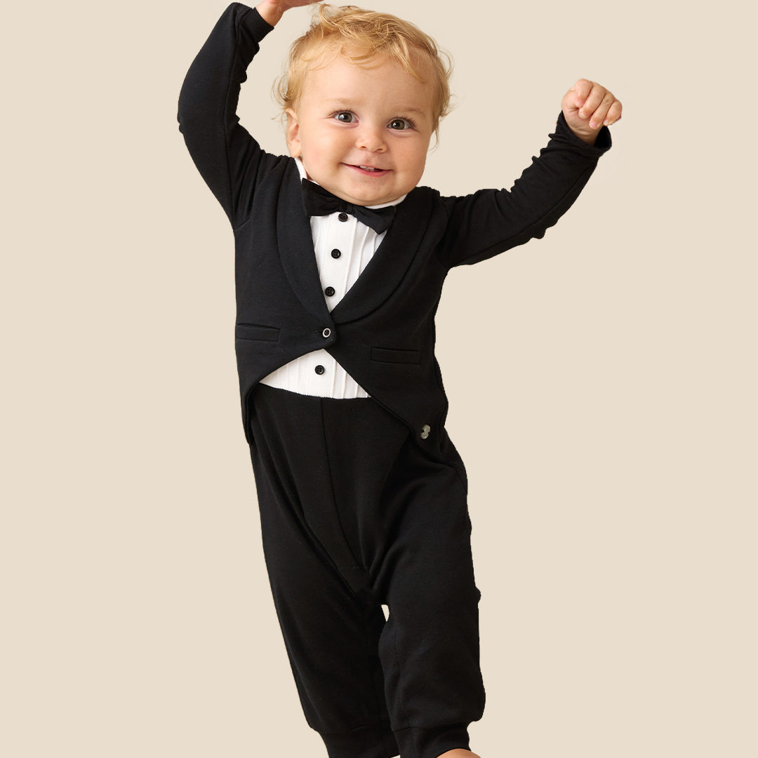 A baby wearing a black formal tuxedo-style romper with a white shirt and black bow tie, posing with arms raised against a plain beige background.