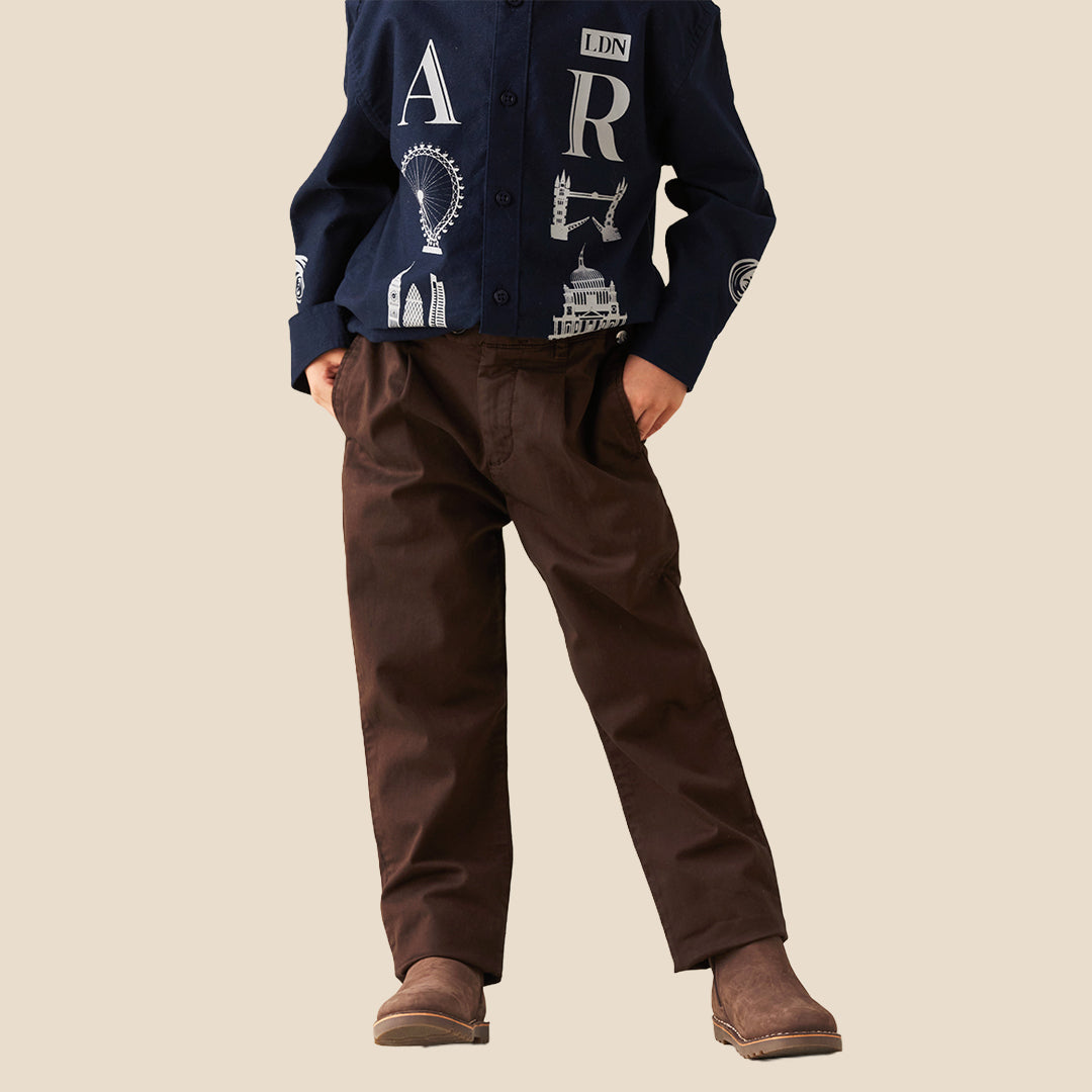 A child wearing dark brown trousers and a navy blue button-up shirt with white graphic prints, standing against a solid beige background.