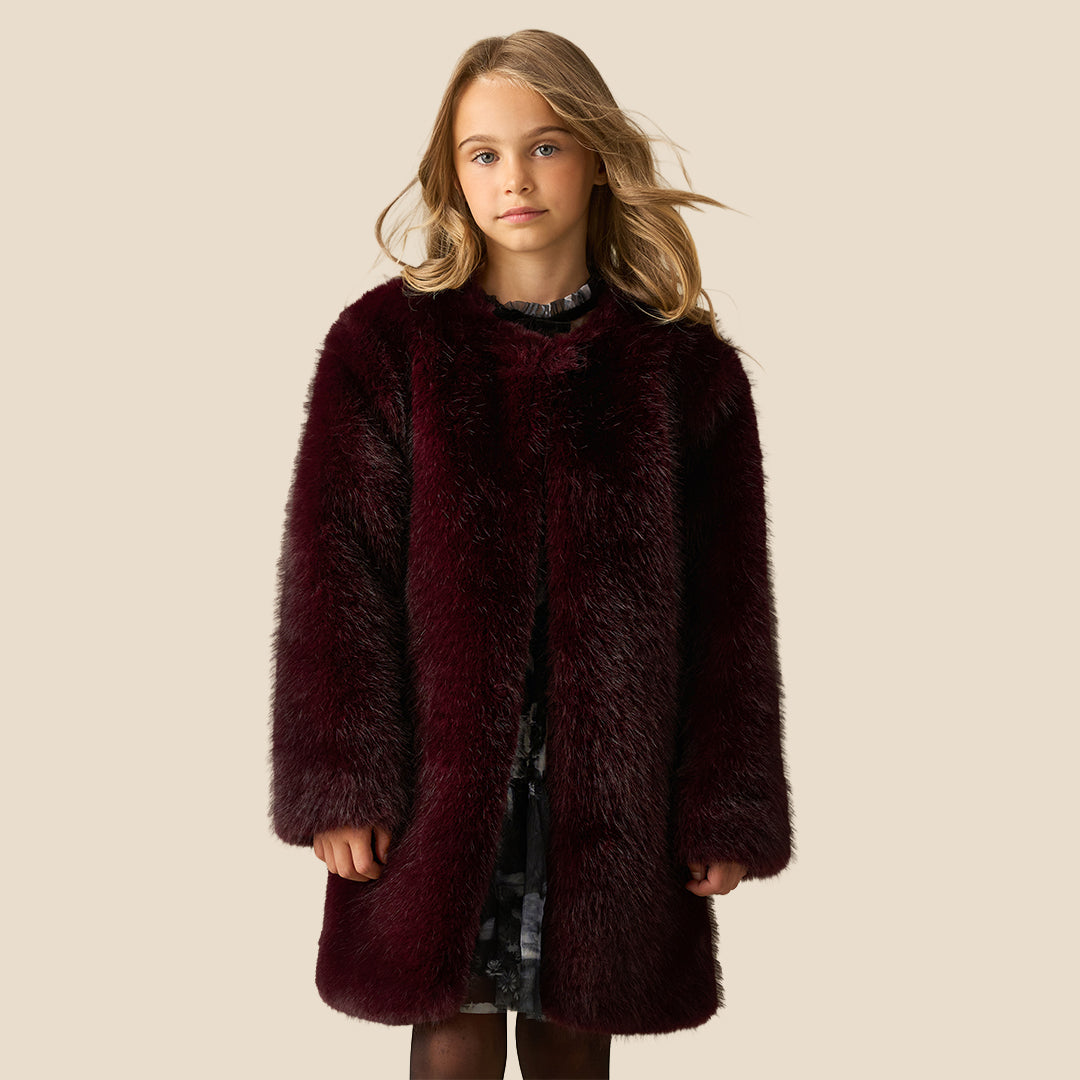 A young girl with blonde hair wearing a plush, dark burgundy faux fur coat over a dark patterned dress, standing against a solid beige background.