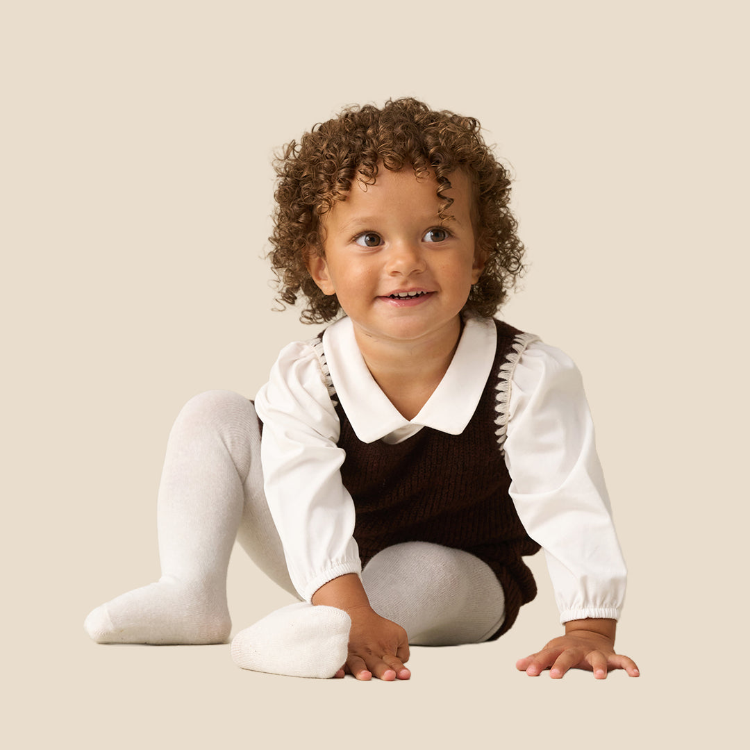 A toddler with curly brown hair wearing a dark brown corduroy pinafore dress over a white long-sleeve collared shirt and white tights, standing against a solid light beige background.