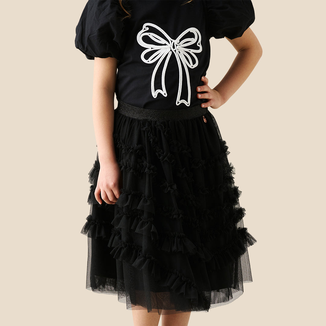 A child wearing a black puff-sleeve top with a large white bow graphic and a layered black ruffled tulle skirt, shown against a plain beige studio background.